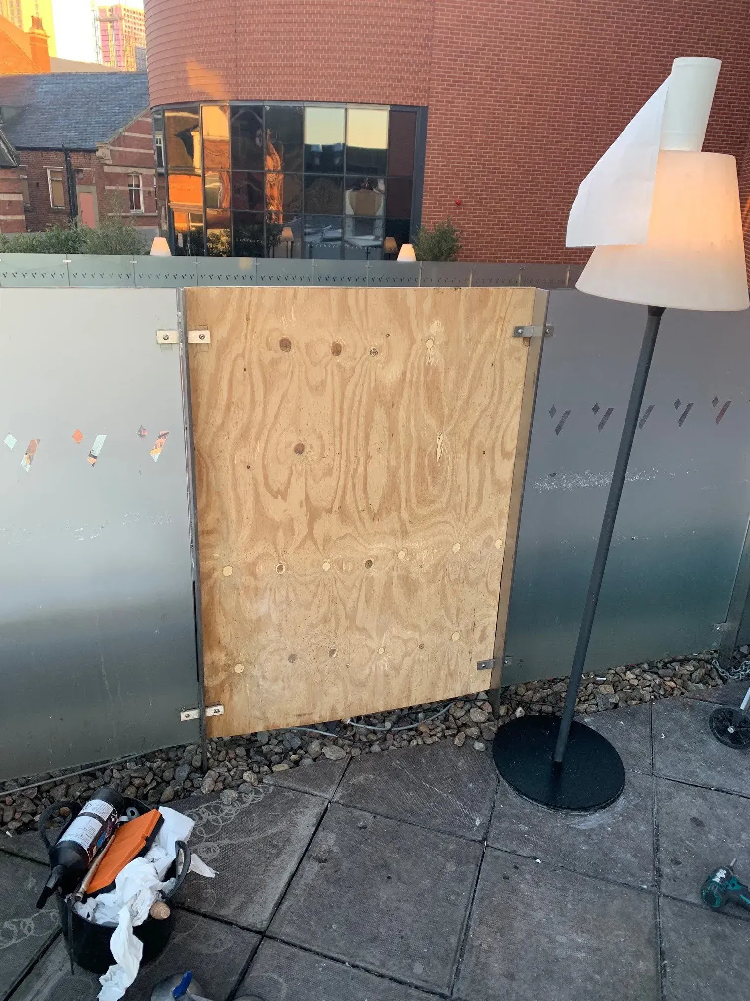 A plywood door is installed in a metal wall. A lamp stands beside it on a stone patio.