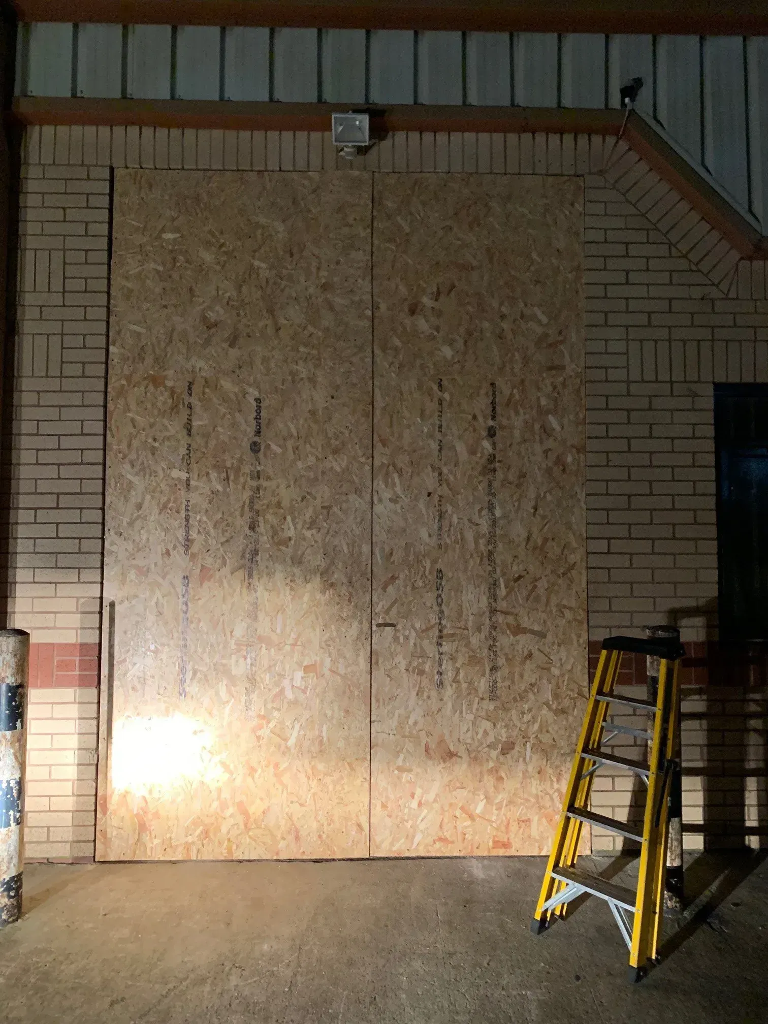 Large plywood boards covering a doorway on a brick building with a yellow ladder propped against the side.