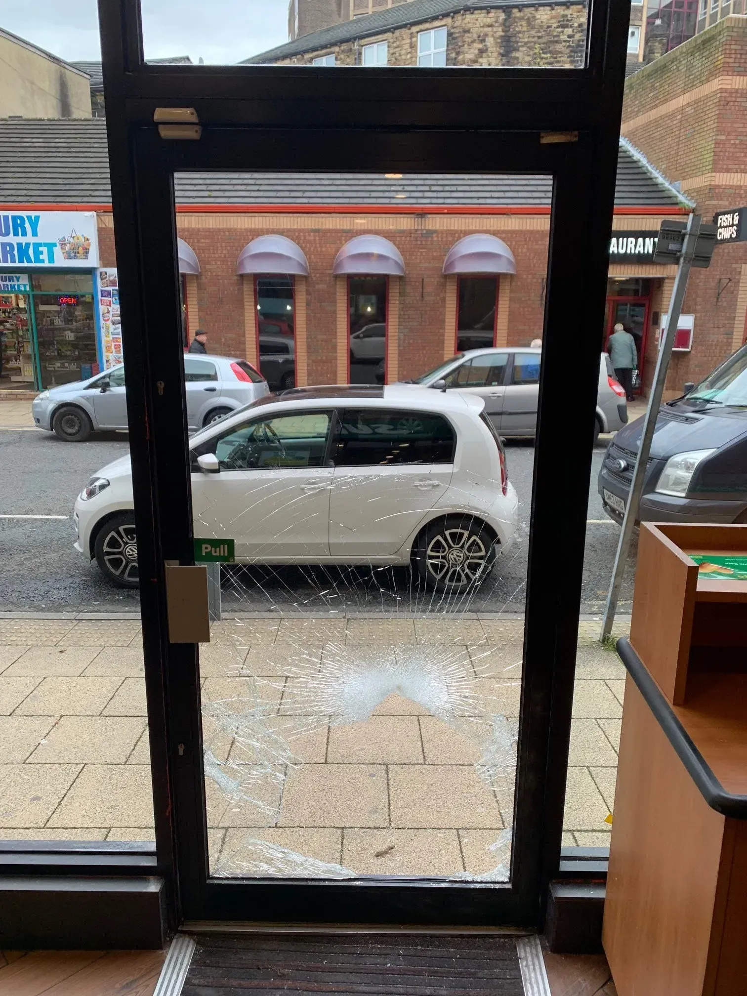 A glass door with a large hole, broken glass on the ground. A white car and street scene visible outside.