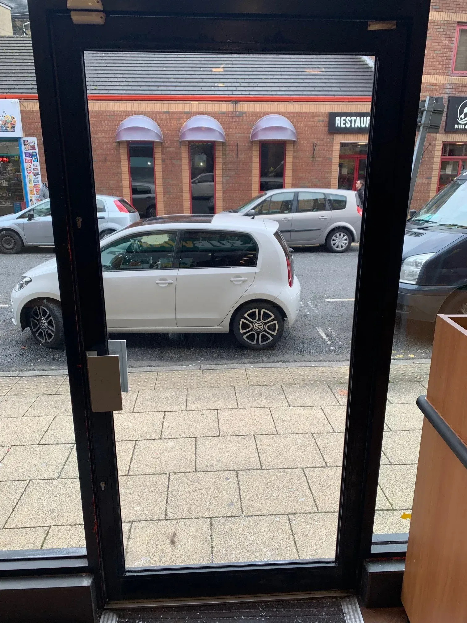 View through a shop doorway: white car parked on street, brick building in background.