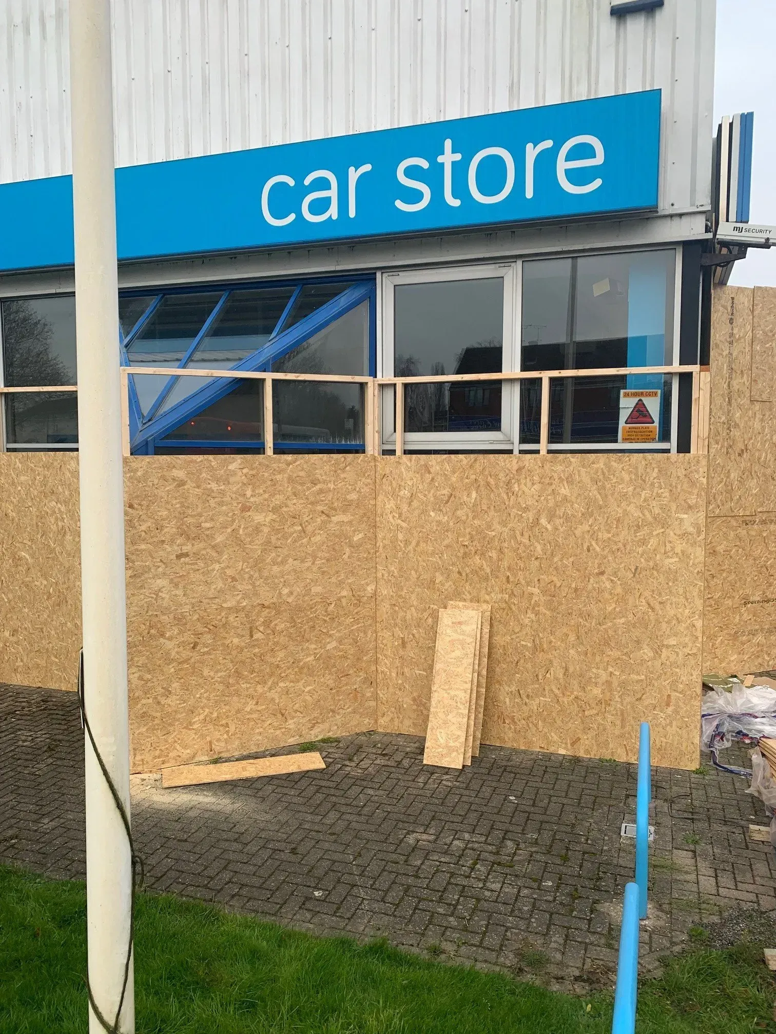 Car store storefront, boarded up with plywood, blue signage.