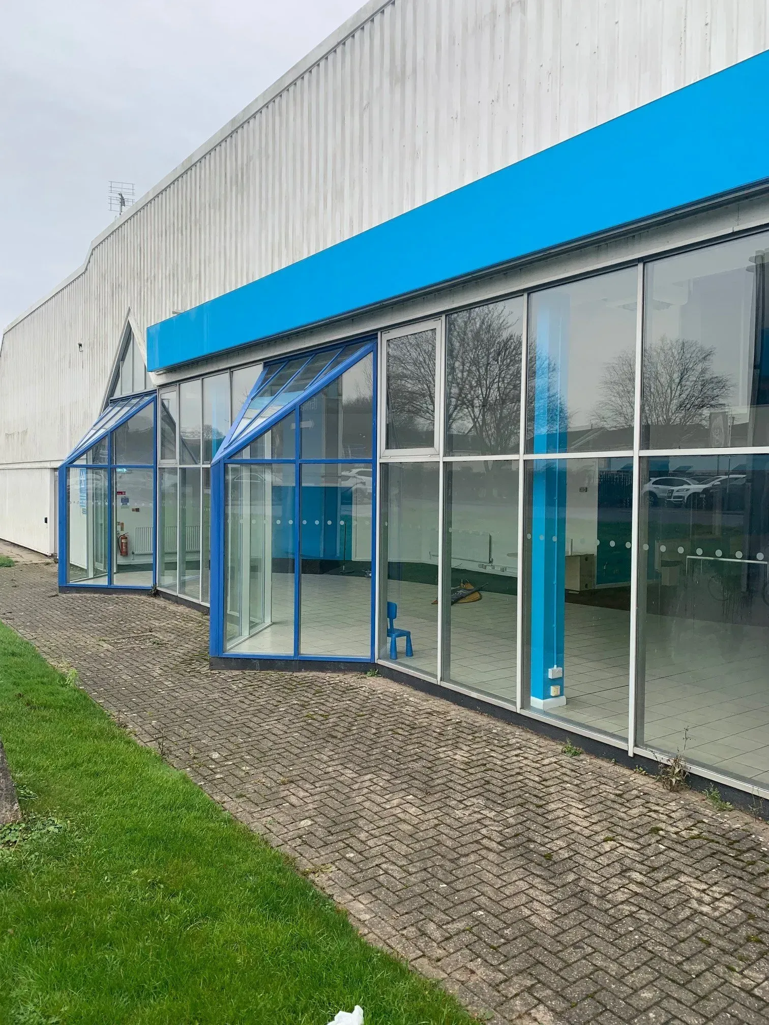 Exterior view of an empty commercial building with blue accents on glass and facade, surrounded by grass and a brick path.