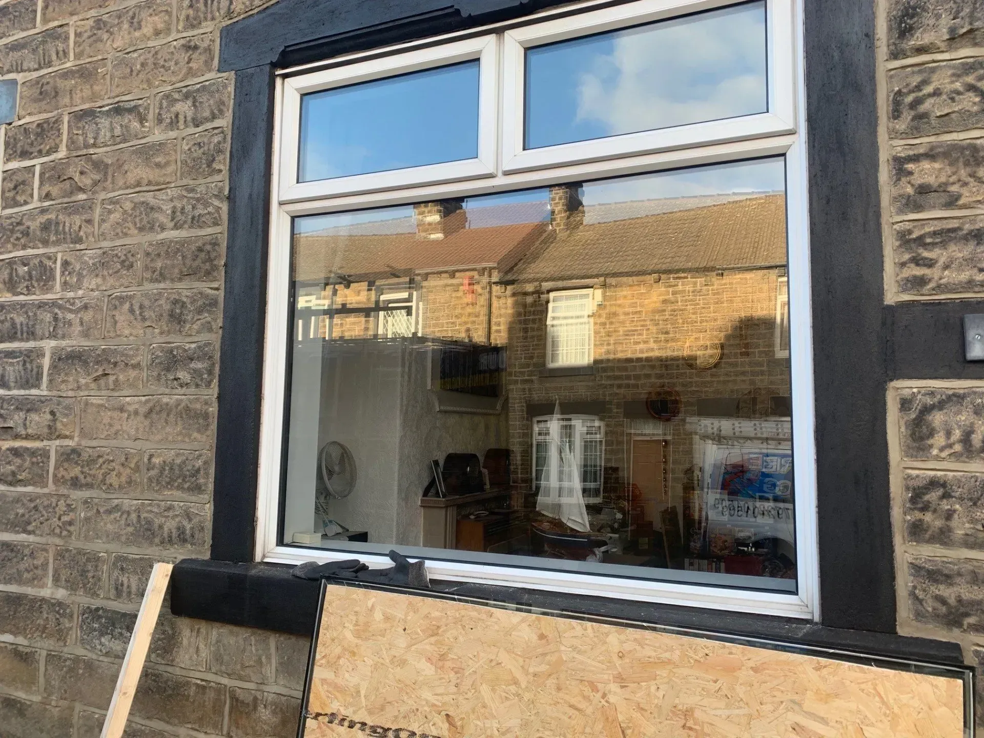 Window framed by black, on a brick building, reflecting buildings and the sky, with plywood beneath.