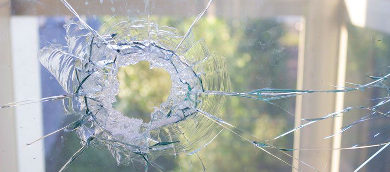 A shattered glass window with a circular hole and radiating cracks. Sunlight and a blurry outdoor view are visible.