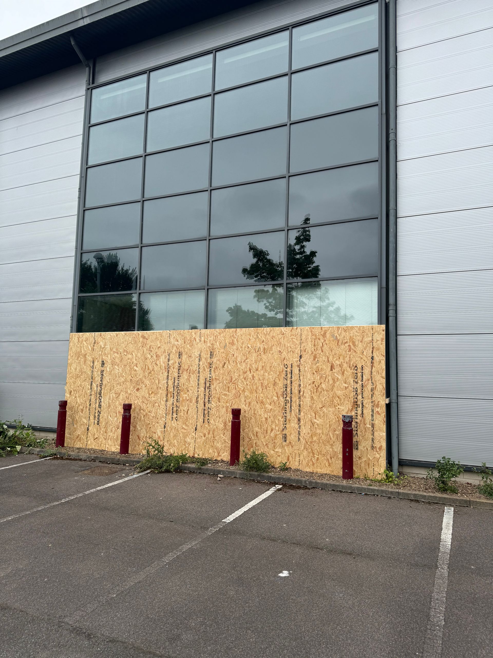 Building with large windows, boarded up with wood. Red bollards in front.