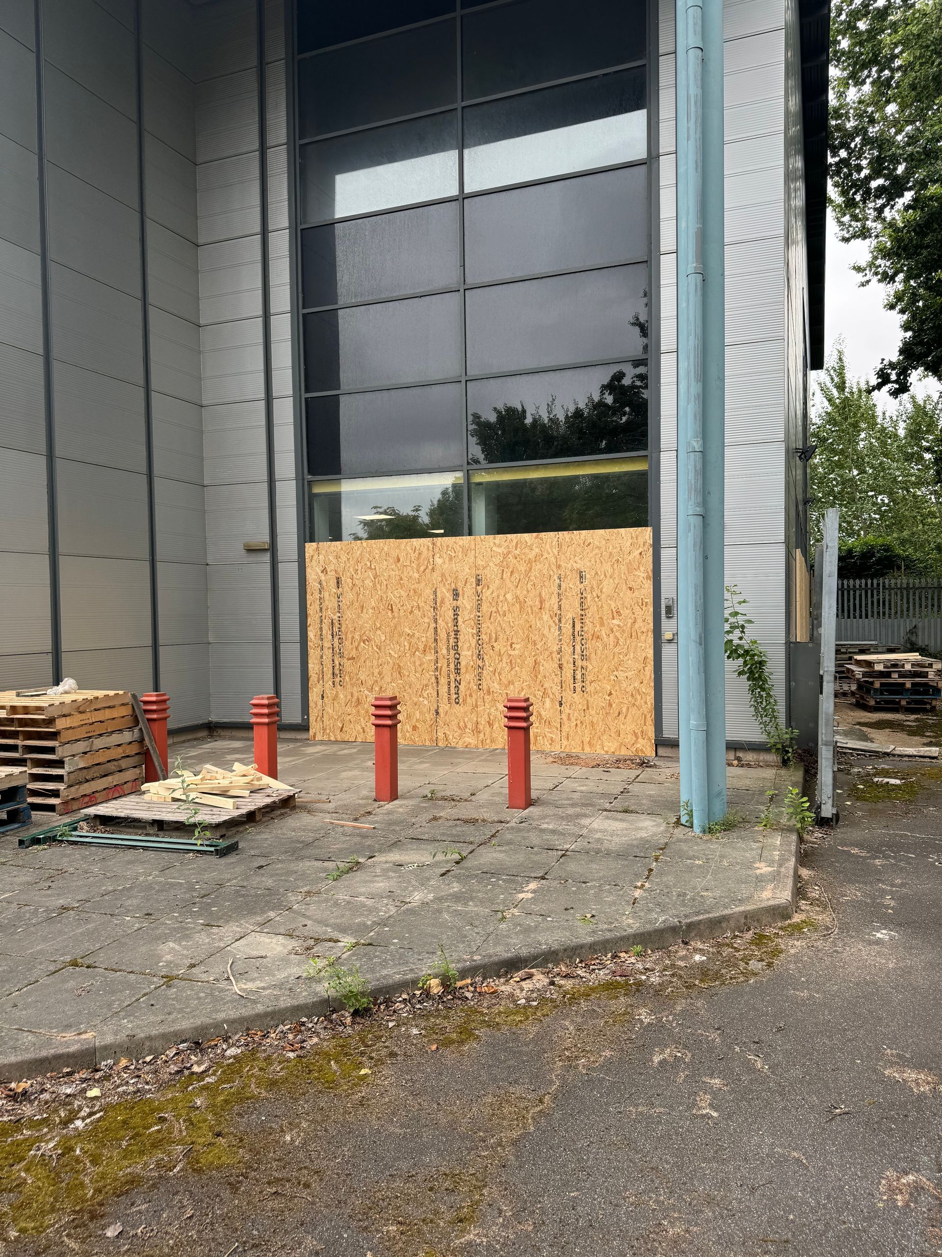Exterior view of a building with boarded-up window, metal siding, concrete ground, red bollards, and pallets.