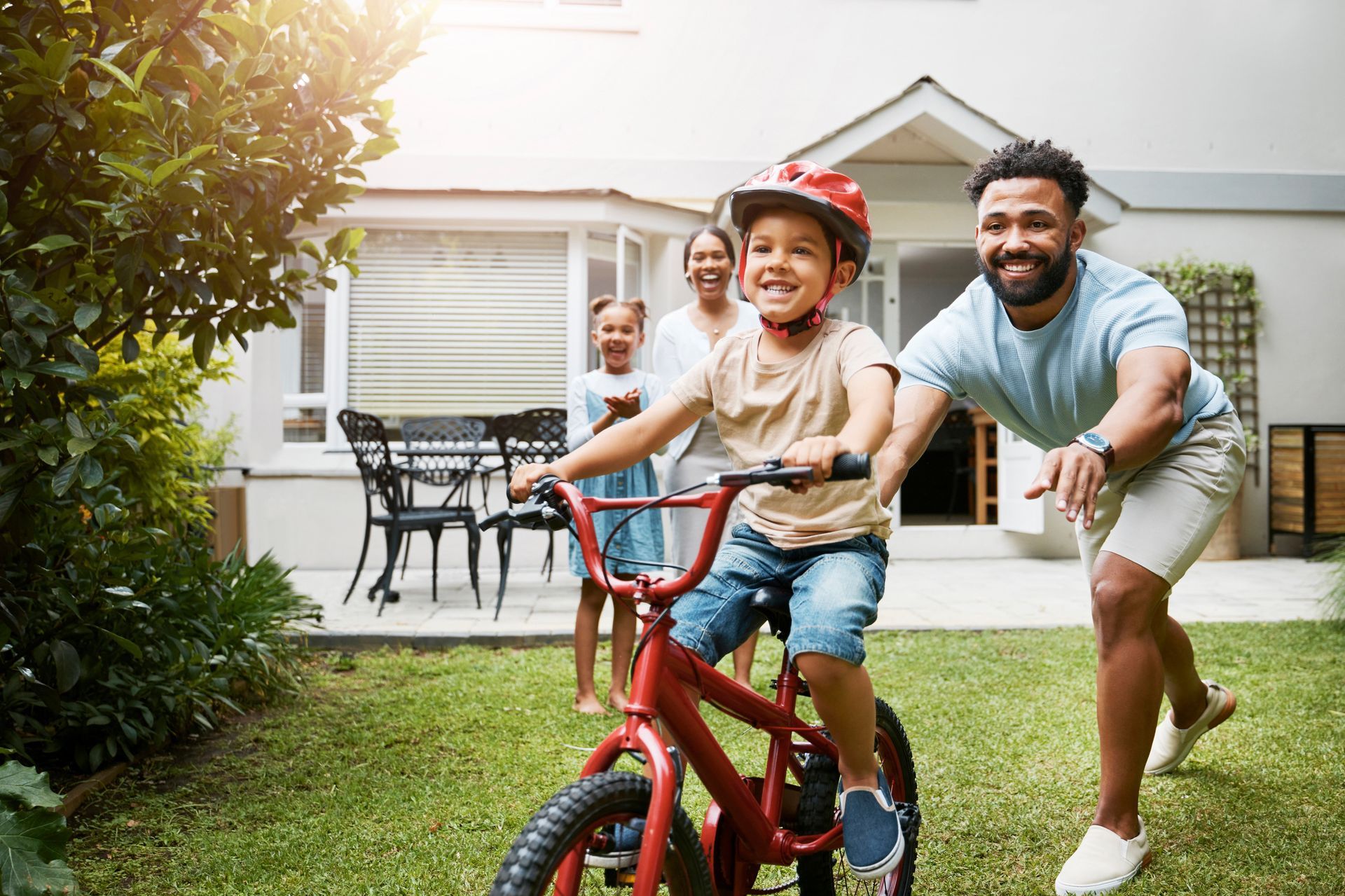 A boy rides a red bicycle in a yard, smiling as his father runs alongside, with a woman and girl watching from the porch.