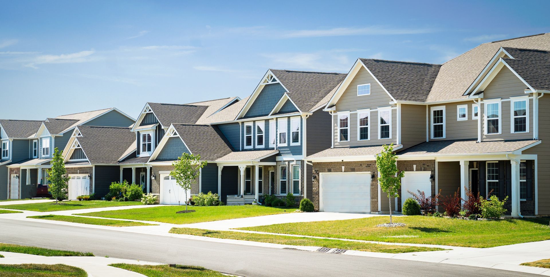 Row of suburban houses with varying colors and garage doors, set against a blue sky. Green lawns and a street are in the foreground.