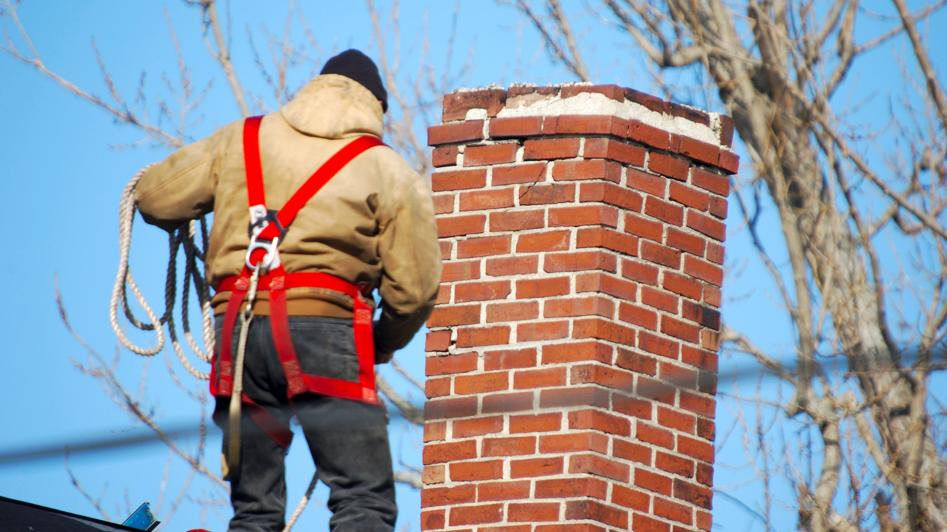 A person wearing safety harness gear stands on a roof next to a brick chimney against a clear blue sky.