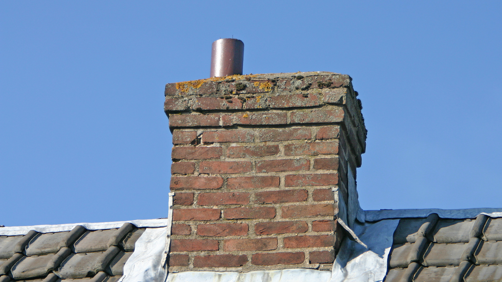 A brick chimney on a roof with a metal flue pipe extending from the top against a clear blue sky.