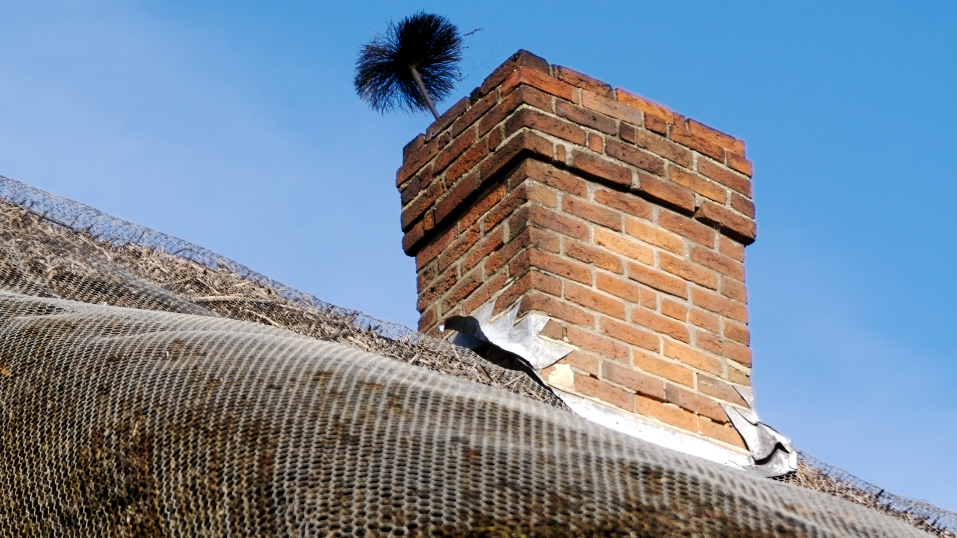 A chimney brush cleaning a brick chimney on a roof with wire mesh netting against a clear blue sky.