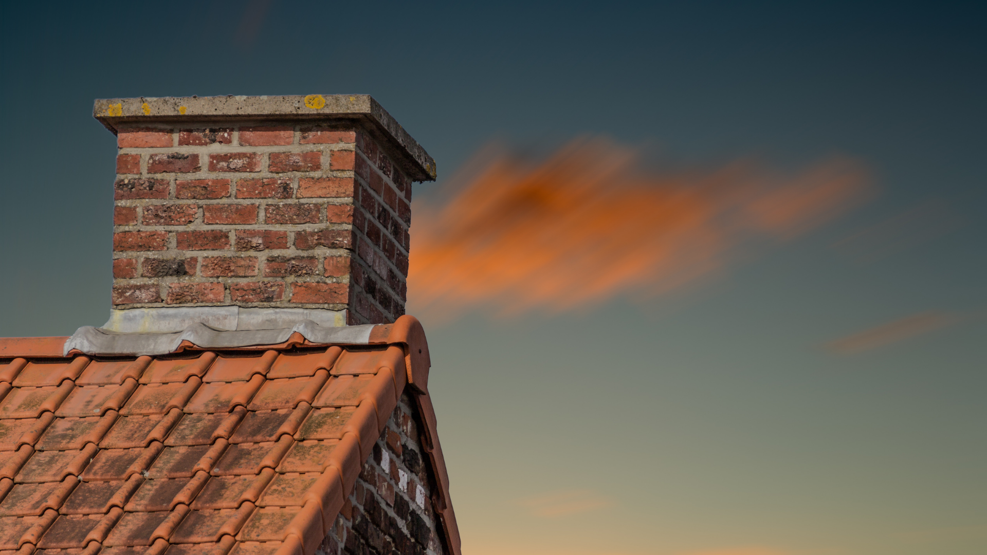 A brick chimney on a red-tiled roof set against a twilight sky with a soft orange cloud.