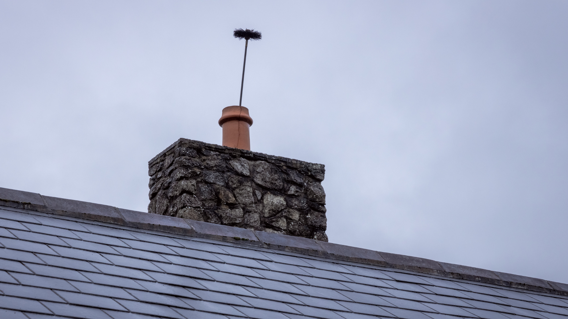 A stone chimney topped with a terracotta pot and a tall, thin metal spike against a gray, overcast sky.