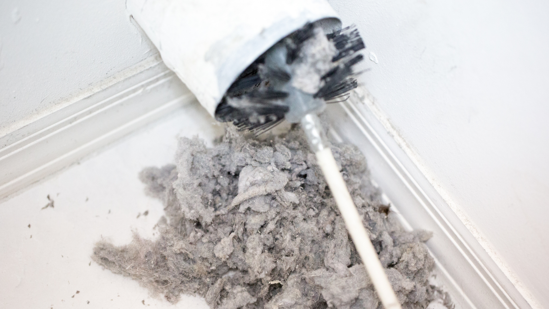 A brush tool pulling a large clump of gray lint and dust out of a white dryer vent pipe against a white wall.
