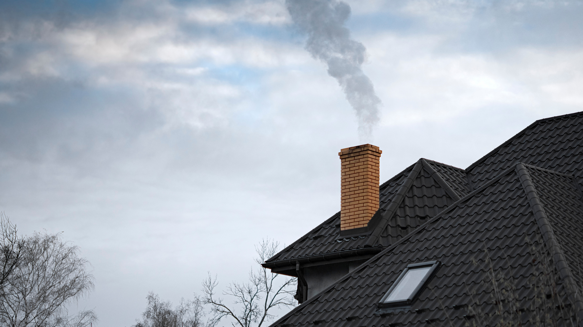 A brick chimney on a dark, tiled roof emits smoke into a cloudy sky.