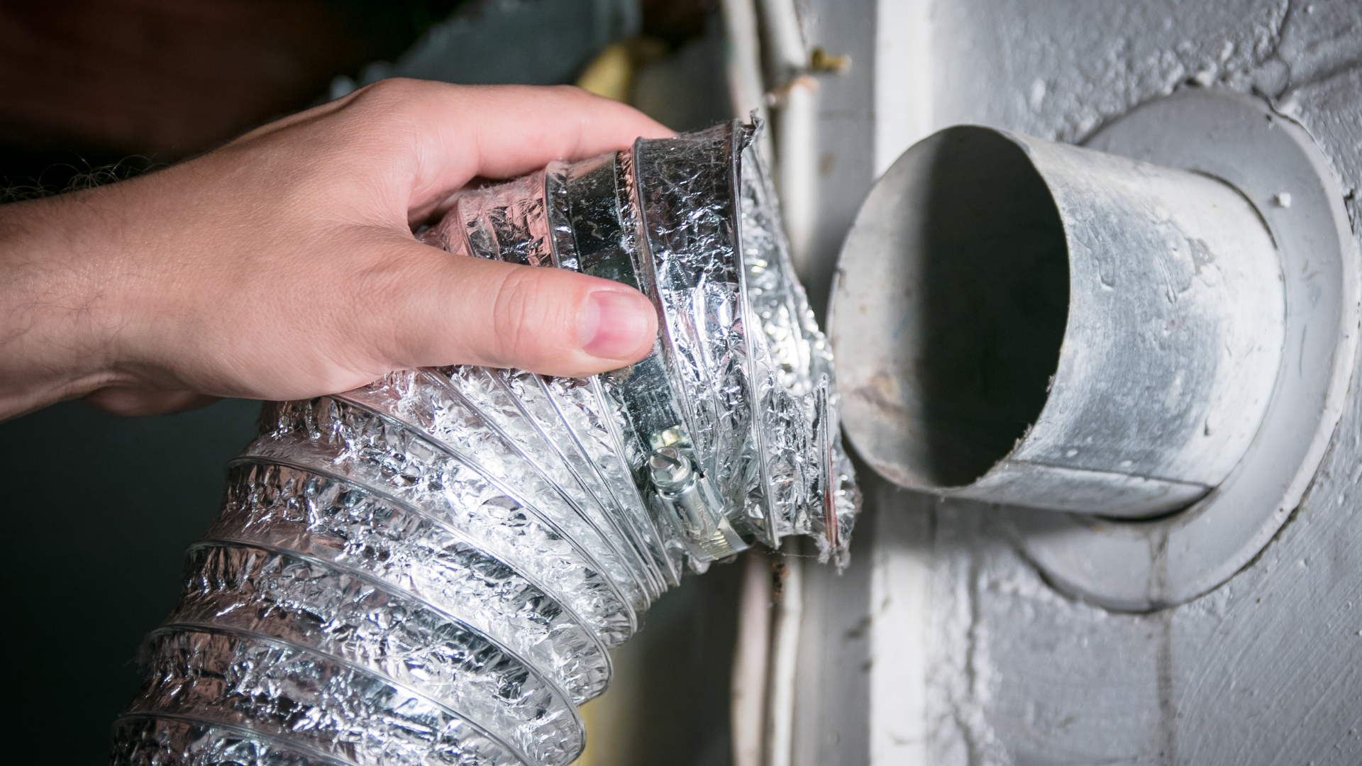 A hand holds a flexible, silver foil dryer vent hose near a wall-mounted metal exhaust pipe.
