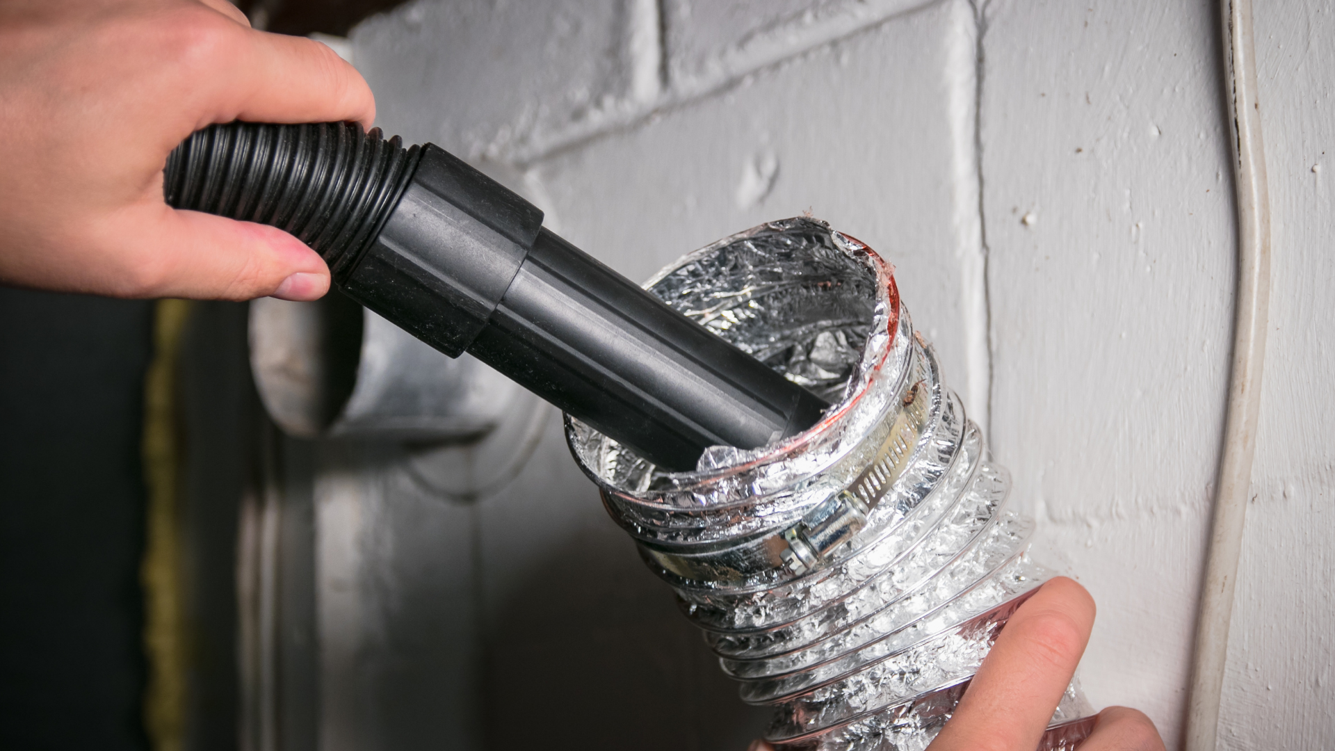A person uses a vacuum attachment to clean the inside of a silver foil dryer vent hose attached to a white brick wall.