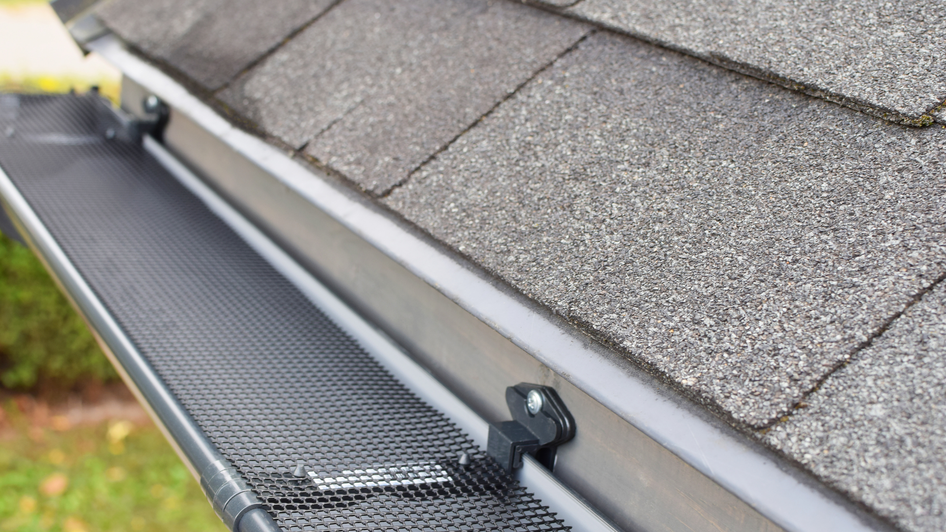 A close-up view of a metal roof gutter with a black mesh guard installed along the edge of grey asphalt shingles.