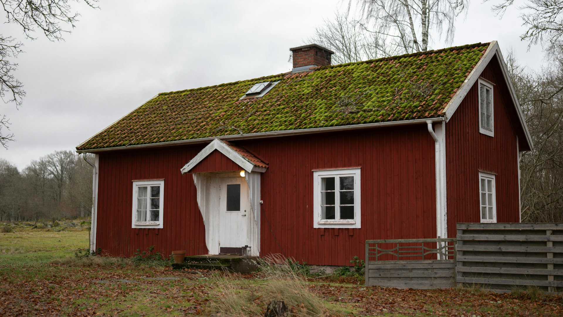 A rustic, red wooden house with a green mossy roof and white trim, set in a field with scattered autumn leaves.