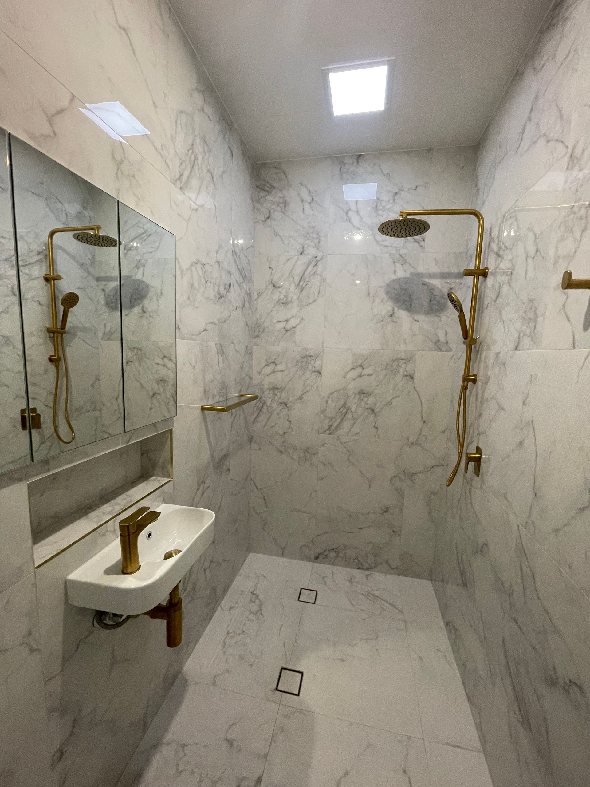 Marble-tiled bathroom with gold fixtures: showerhead, faucet, towel bars. Includes a sink, mirror, and recessed shelf — Smooth Flow Plumbing Cairns in Cairns City, QLD 