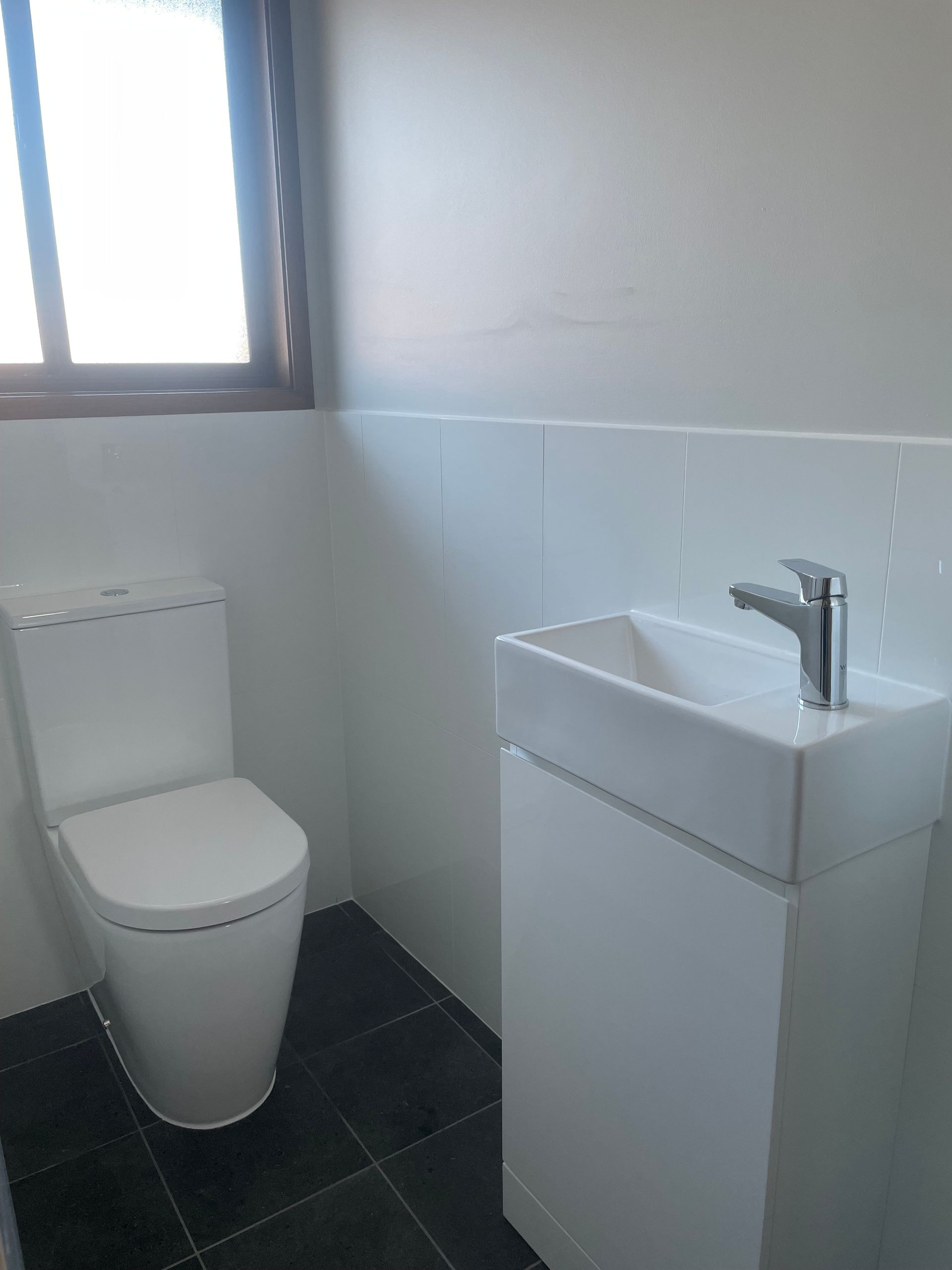 White toilet and sink in a small bathroom with dark tile floor, window, and white walls — Smooth Flow Plumbing Cairns in Cairns City, QLD 