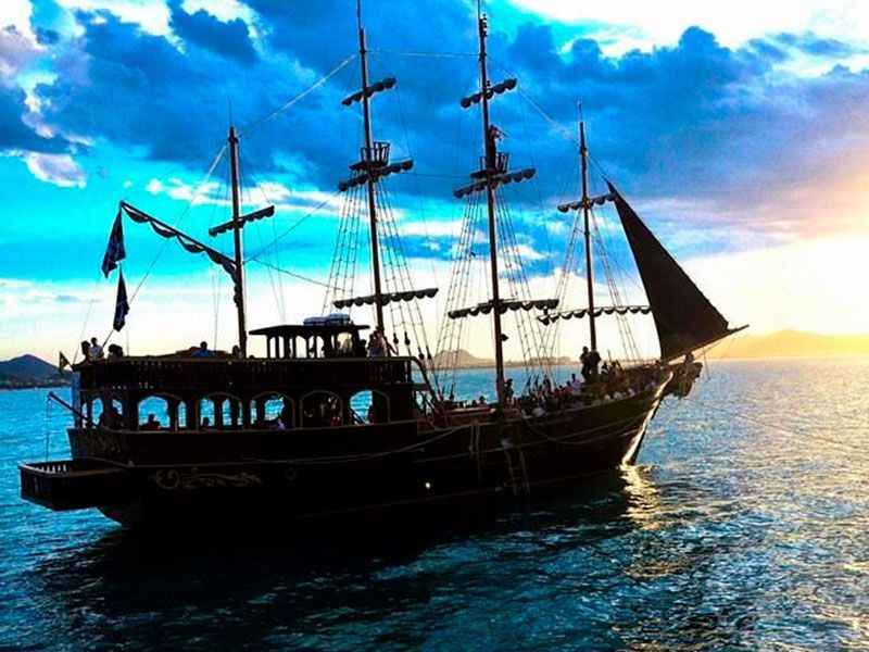 Navio pirata navegando na água, passageiros no convés, pôr do sol ao fundo.