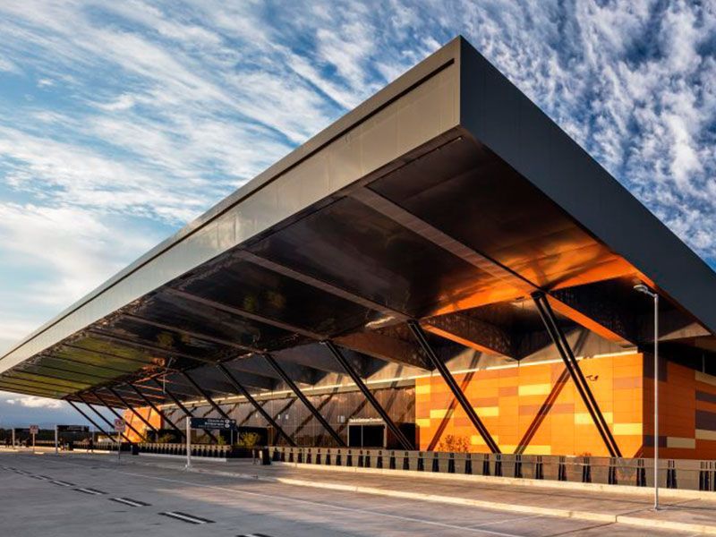 Prédio industrial moderno com uma grande cobertura cinza-escura, painéis laranja e marrons, contra um céu parcialmente nublado.