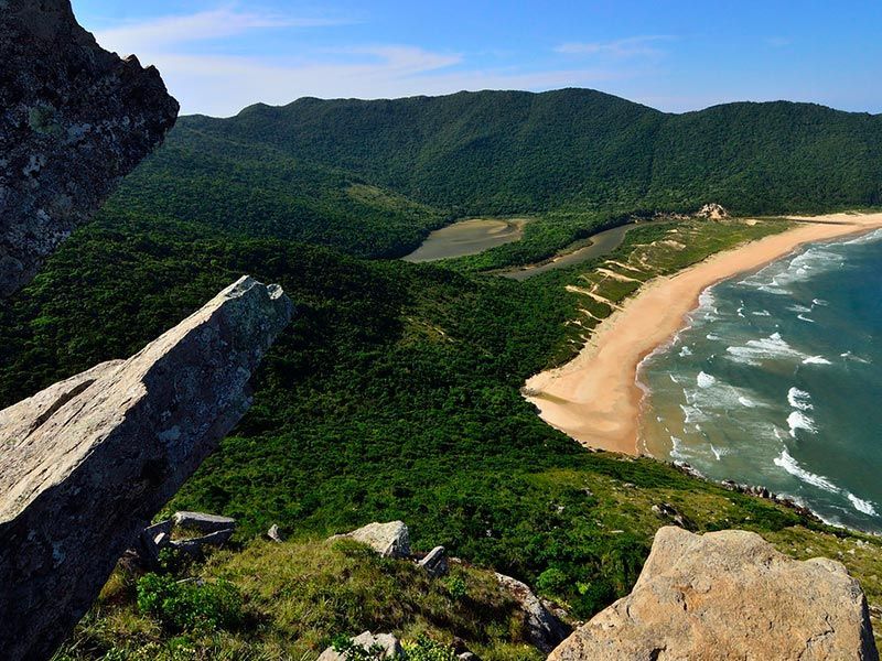 Vista panorâmica: praia costeira com montanhas, árvores verdejantes e água azul sob um céu límpido.