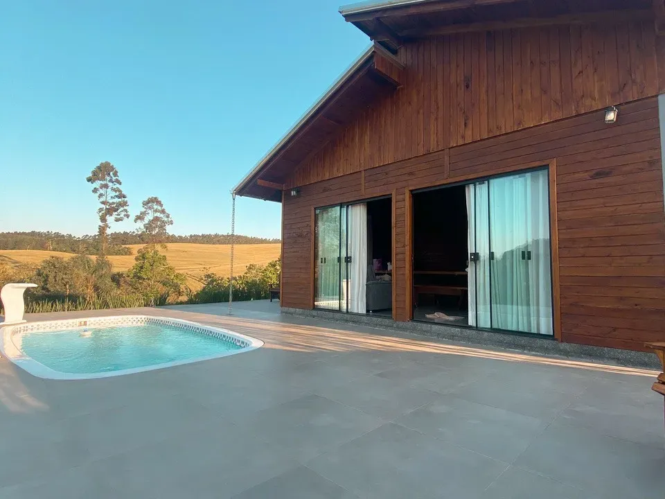 Cabana de madeira com portas de vidro deslizantes, uma pequena piscina e um campo sob um céu azul.