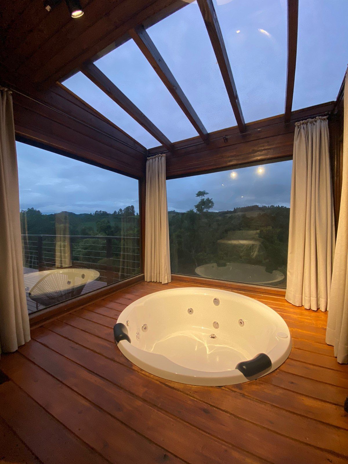 Banheira de hidromassagem interna com deck de madeira, teto de vidro e cortinas, com vista panorâmica para o exterior.