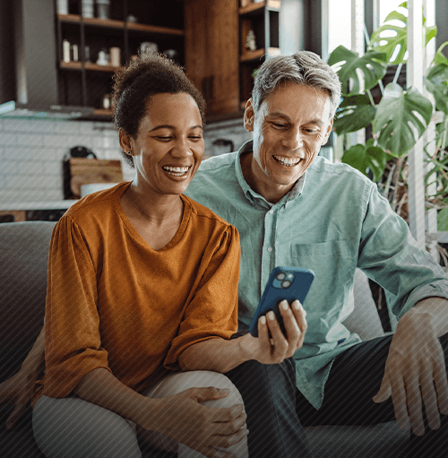 Couple smiling while looking at a phone together on a couch.