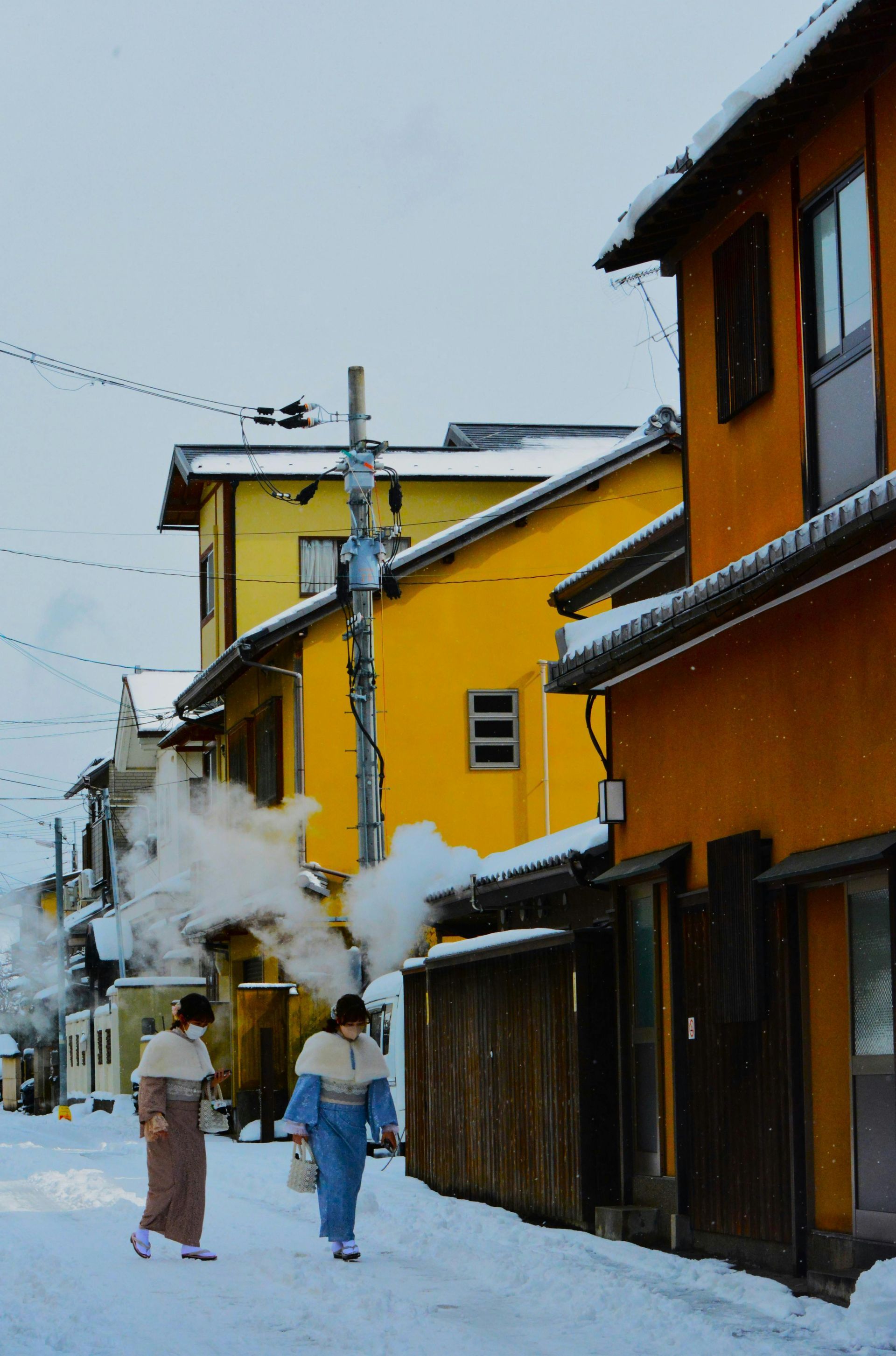 Snowy street in Japan with two people in kimonos walking past colorful buildings. Steam rises.