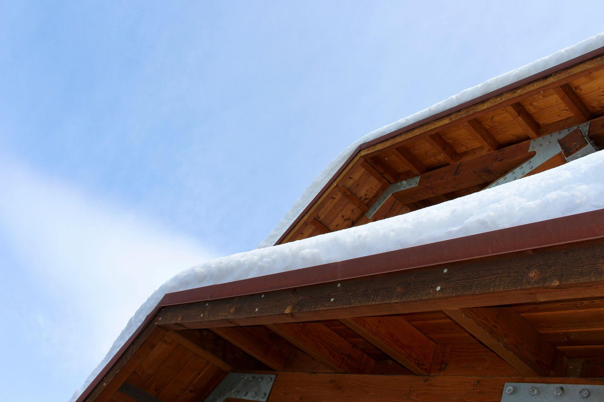 Snow-covered wooden roof against a blue sky. Brown wood and white snow contrast with the bright background.