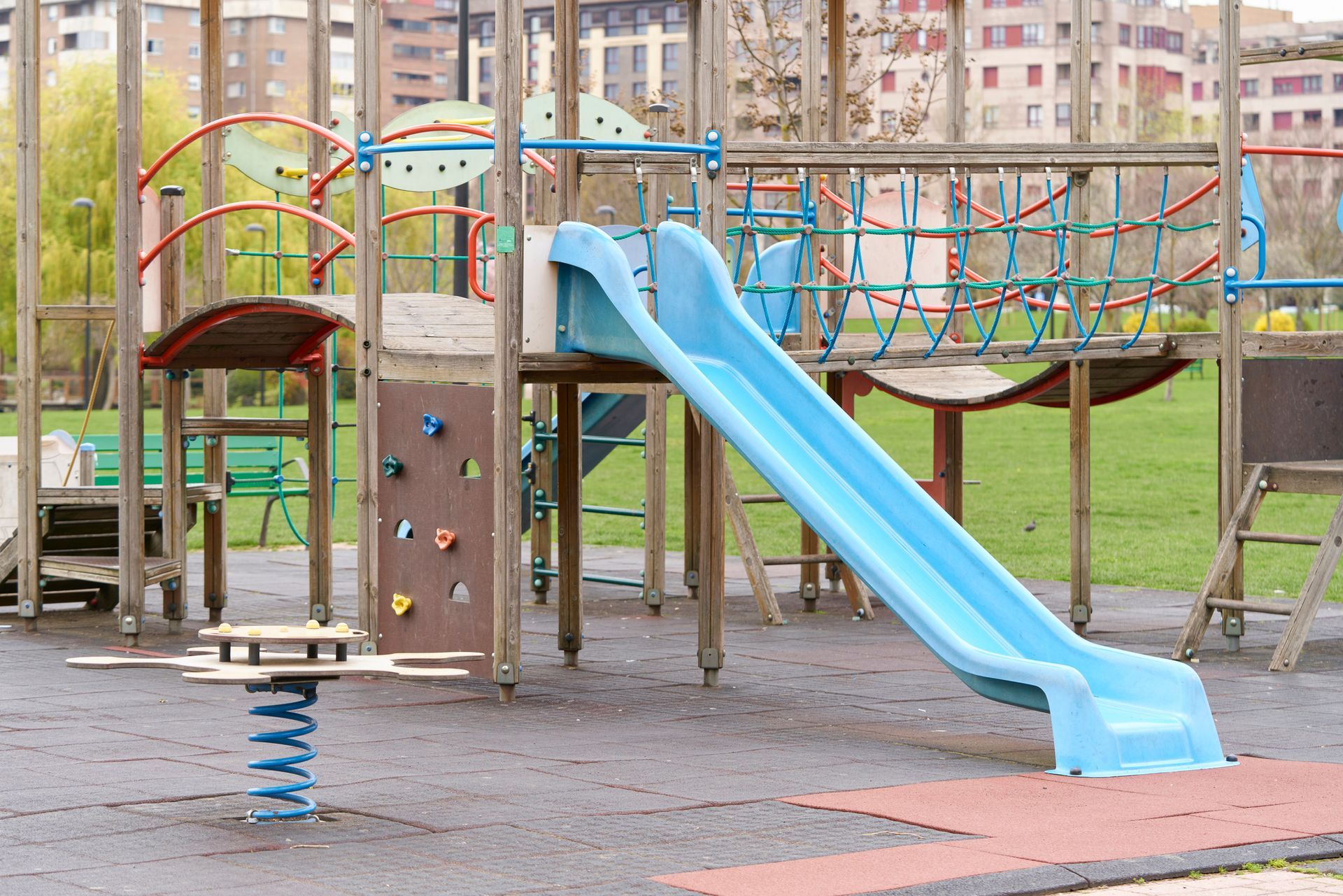 A playground with a blue slide, climbing features, and a ride-on toy set on grass near buildings.