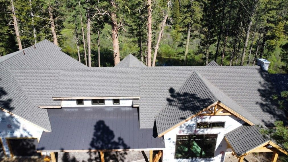 Gray-roofed house with multiple gables and a dark, flat metal roof section; surrounded by trees.