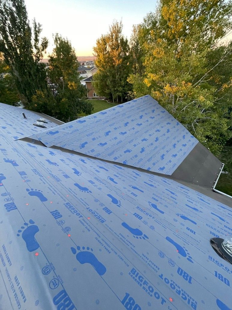 Blue roofing underlayment on a house roof, with trees and a lake in the background.