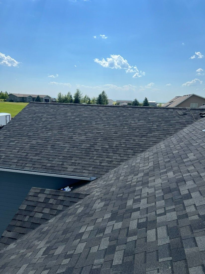 Dark shingled roof under a blue sky, partially obscuring the side of a house.