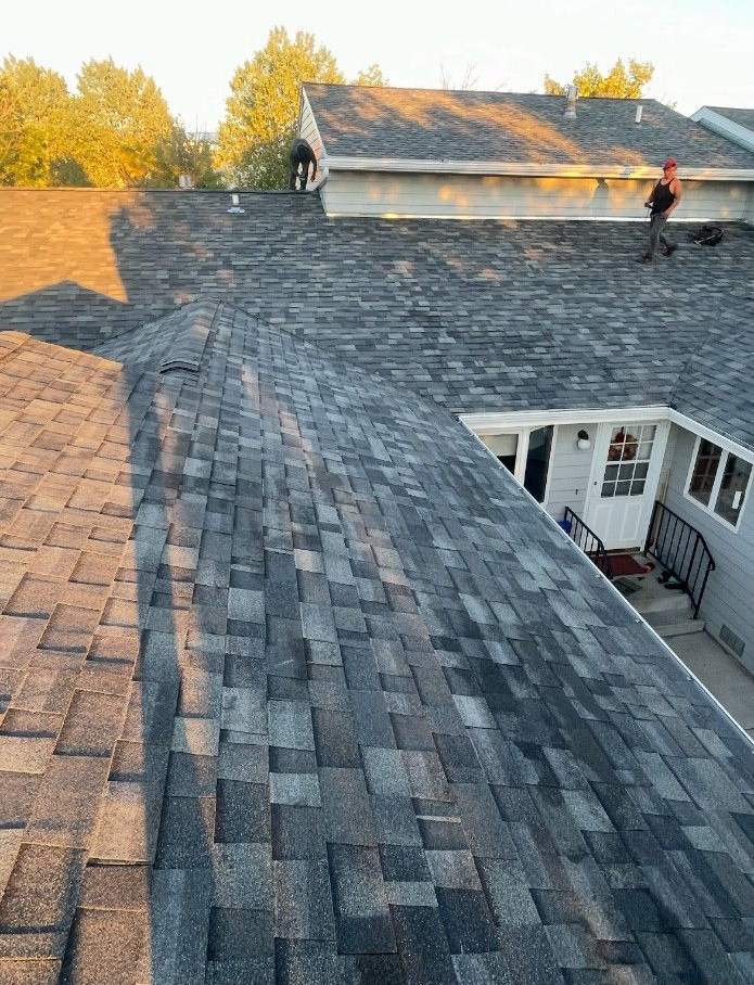 Workers on a roof with gray shingles; sunlight on the roof. Two people. White building and trees in the background.