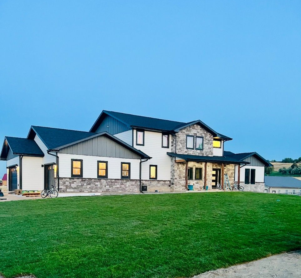 Two-story house with green lawn, stone and white siding, and dark roof under a blue sky.