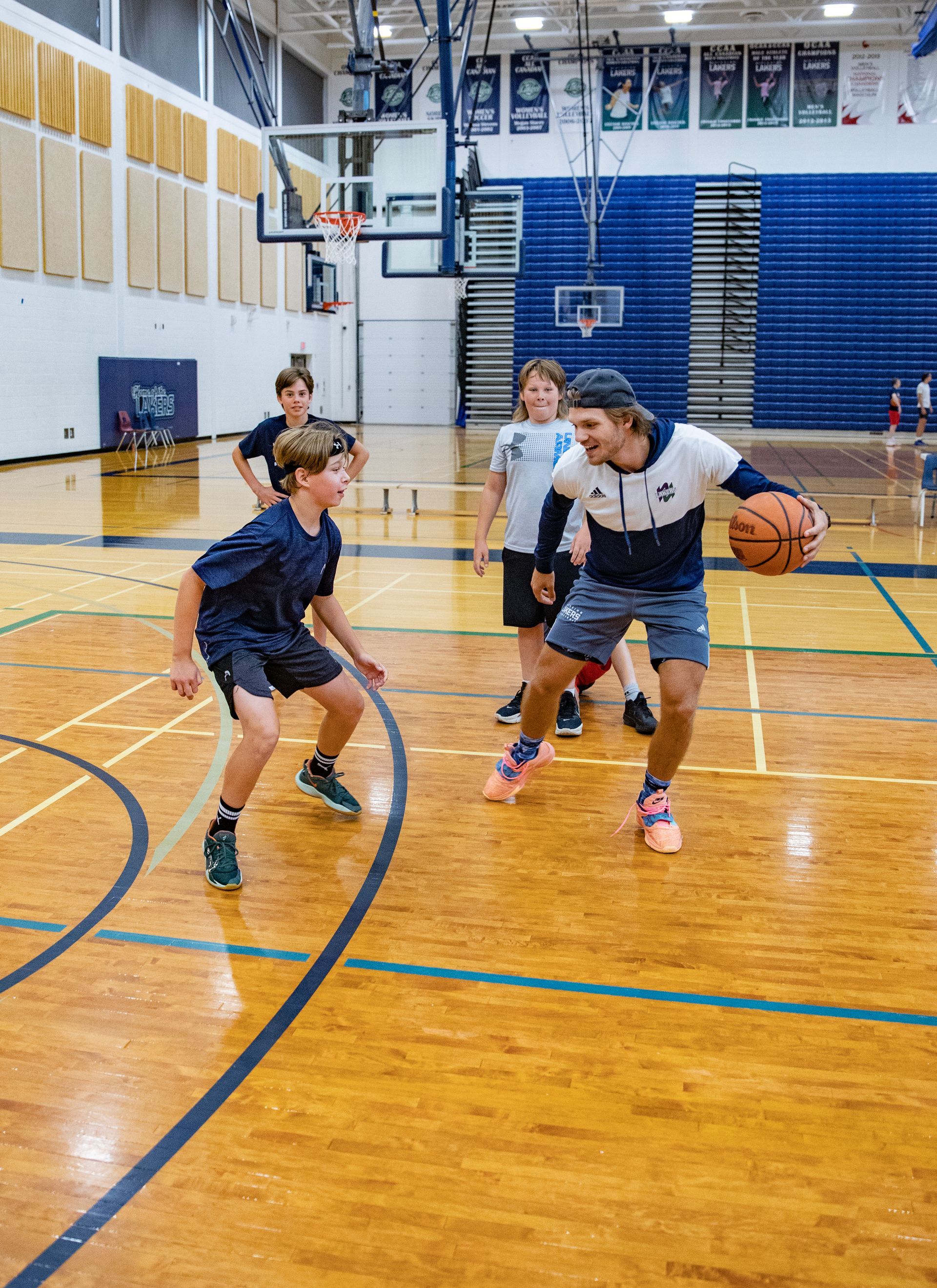 Basketball coach dribbles the ball, facing a young player on a court with other kids in the background.