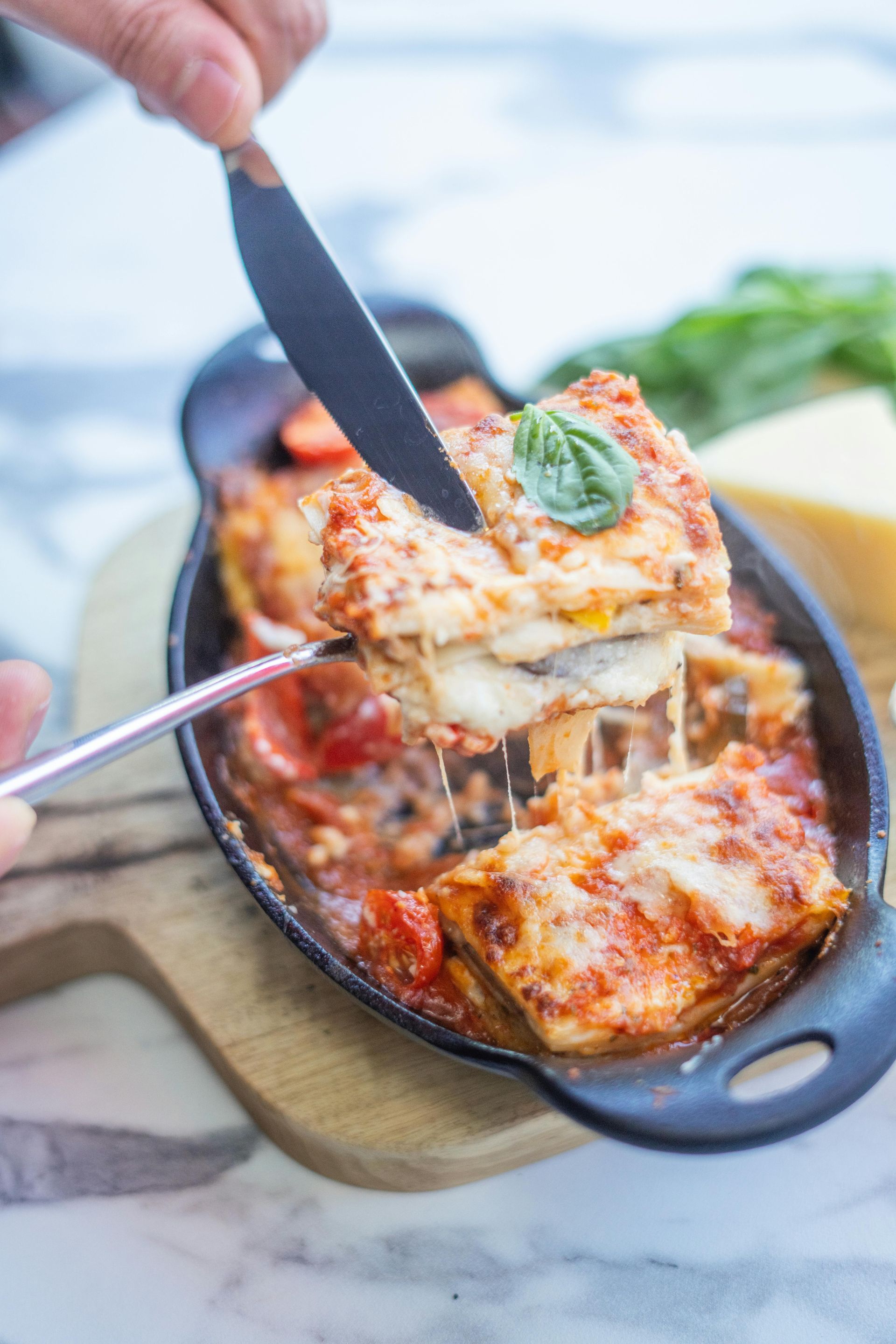 Person cutting cheesy lasagna with a knife and fork in a black dish.