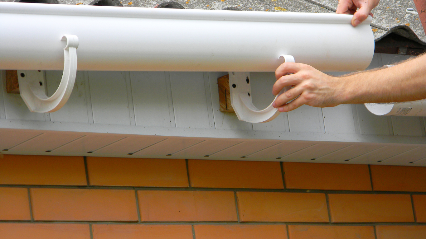 A man is installing a gutter on the side of a brick building.