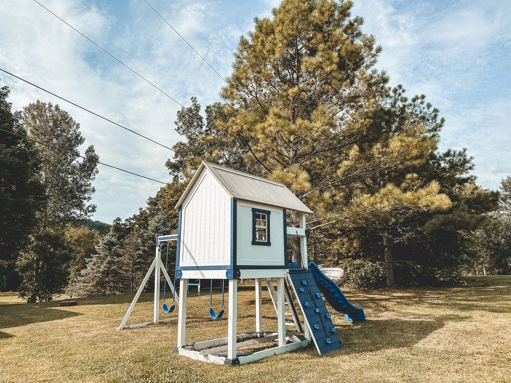 A white and blue tree house with a slide and swings in a yard.