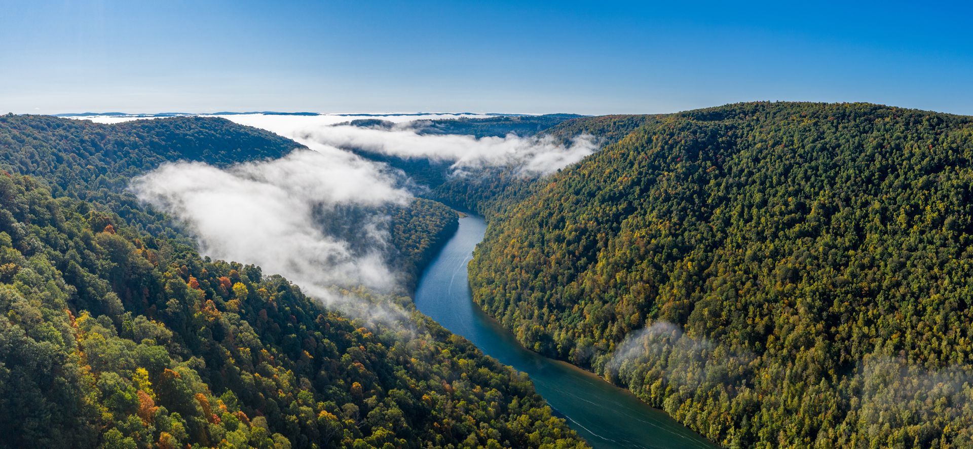 River winds through a valley with forested hills under a blue sky. Mist hangs in the valley.
