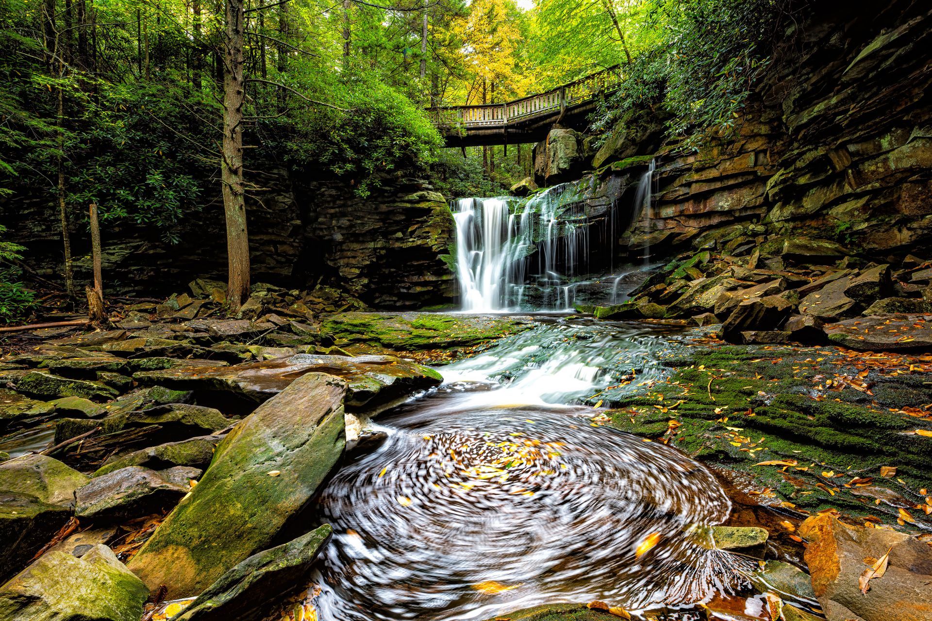 Waterfall cascading into a rocky stream in lush forest, wooden bridge in background.