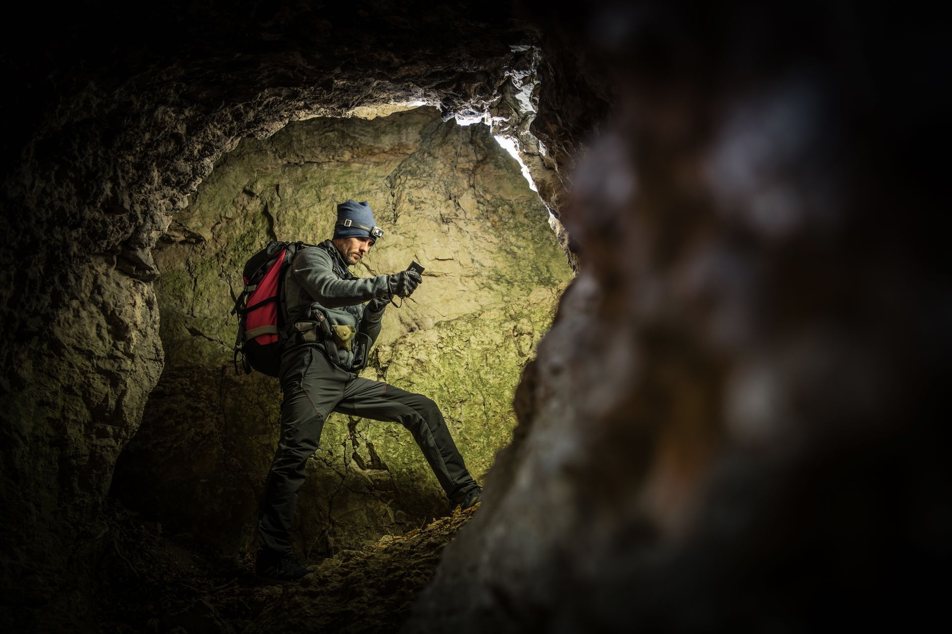 Person in cave with headlamp and red backpack, exploring a rocky passage.