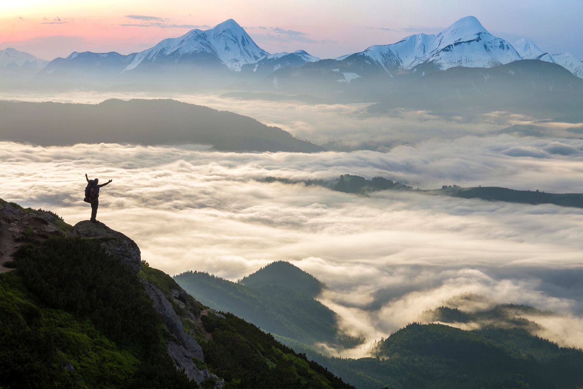 Person with arms raised on a mountain peak, overlooking a cloud-filled valley and snowy mountain range at sunrise.