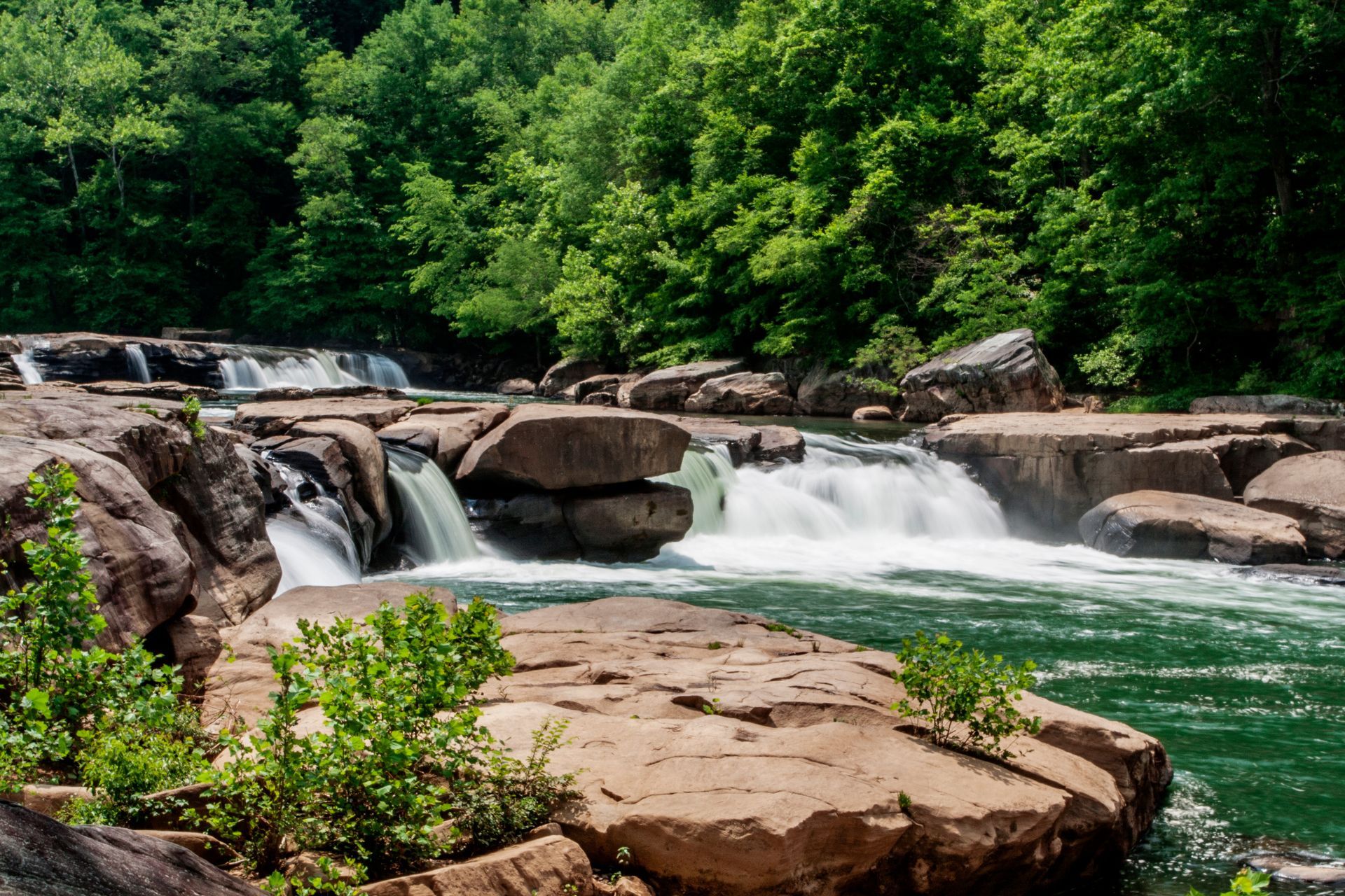Waterfall cascading over brown rocks into a teal river, surrounded by green trees.