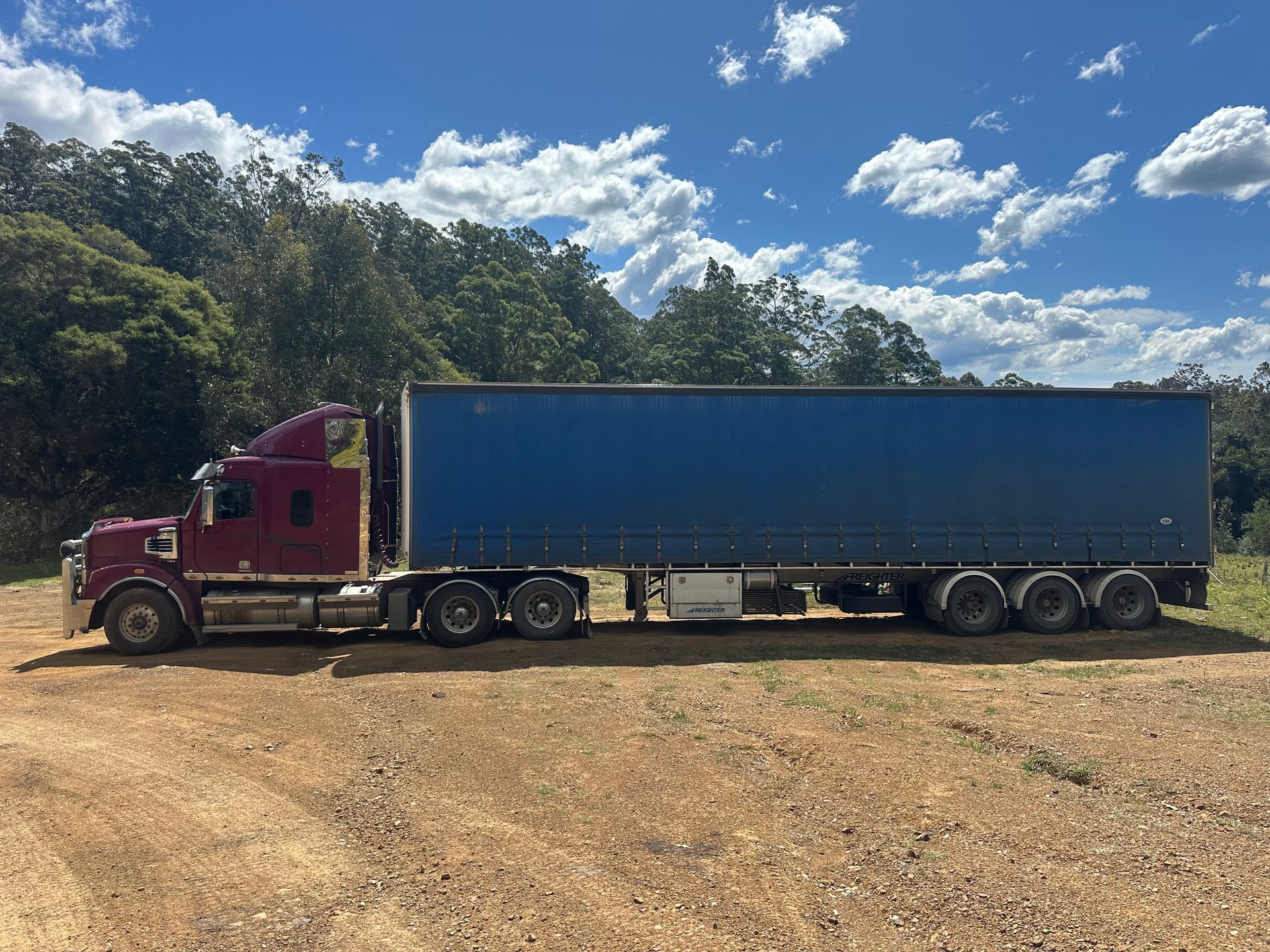 Burgundy freighter truck with blue trailer parked on dirt road under a partly cloudy sky — D & M Freighters Water In Bucca Wauka, NSW