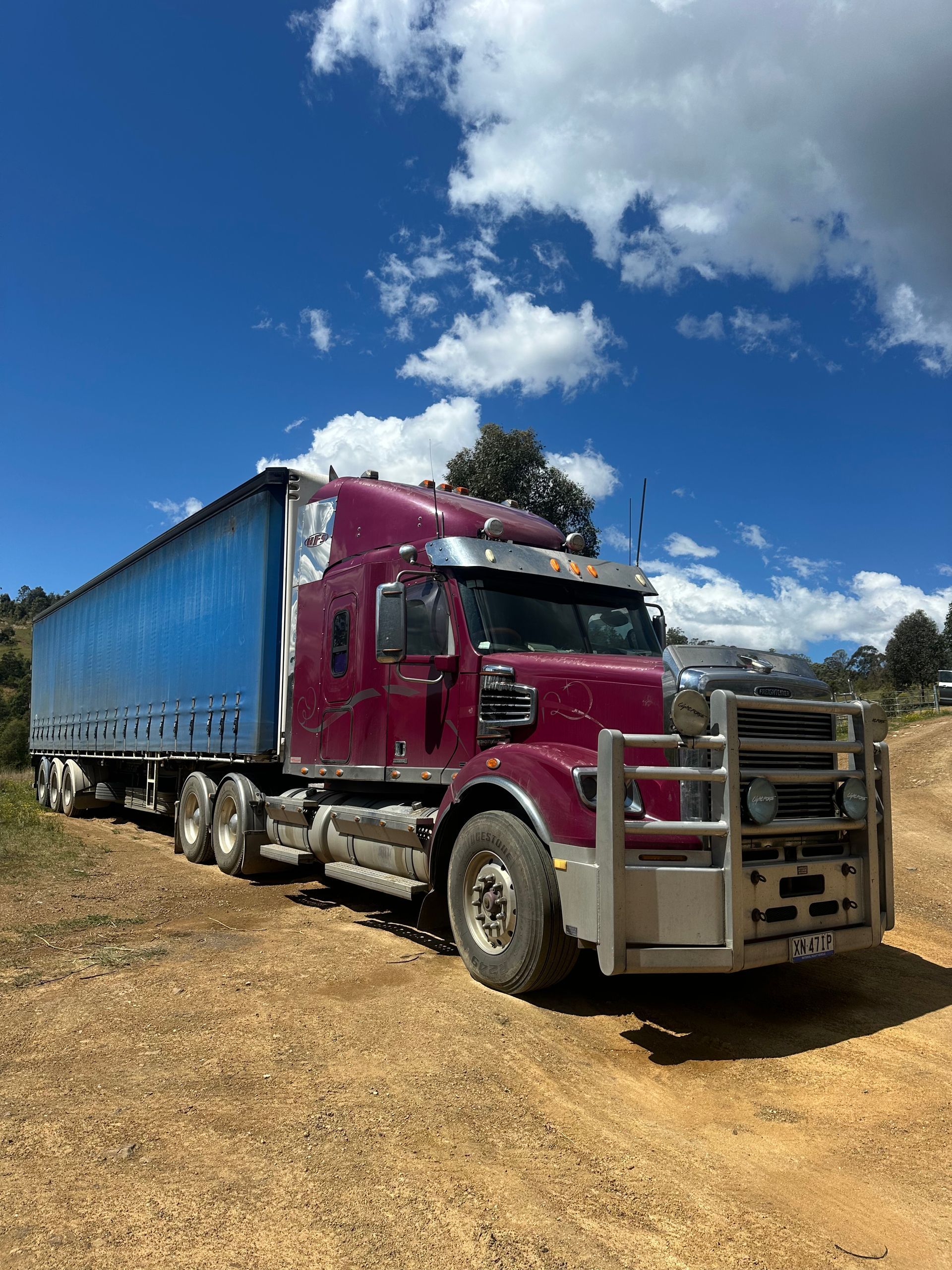 side view of maroon semi-truck with a blue trailer parked on a dirt road under a partly cloudy blue sky — D & M Freighters Water In Bucca Wauka, NSW